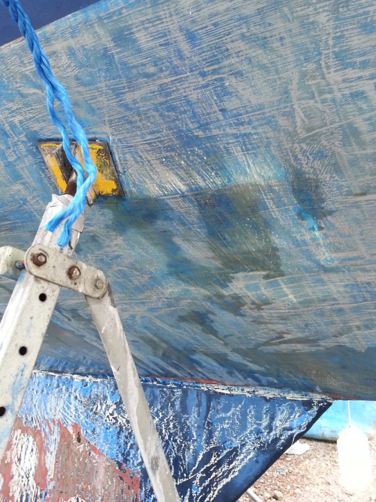 Efter flera omgångar kaustik soda tar vi bort resten med en färgskrapa. Den gråa epoxy grundfärgen framträder tydligt. Ojämnheter i ytan gör att gammal bottenfärg (blå) är kvar.